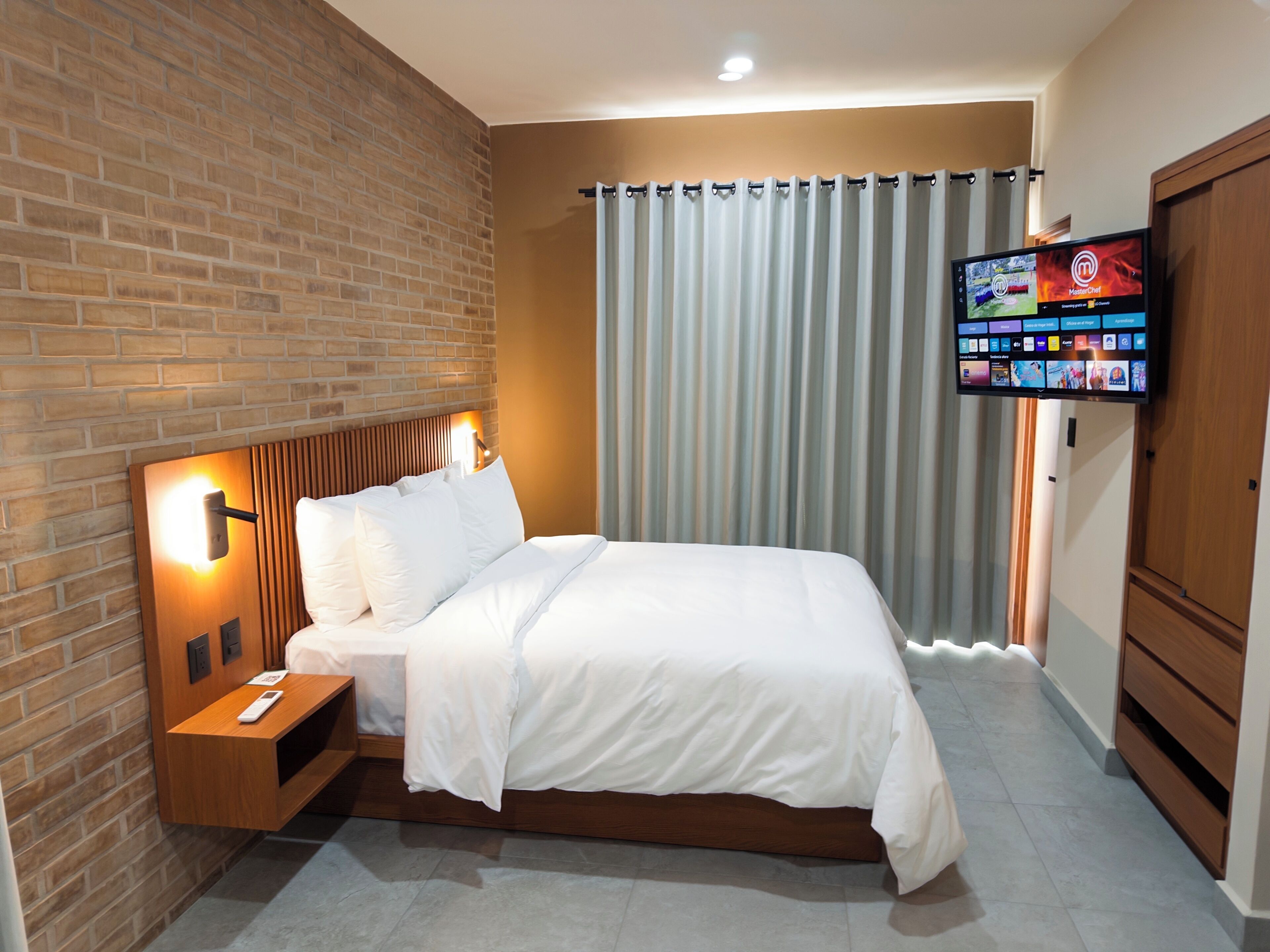 A hotel room with a brick wall, a bed with white bedding, a nightstand with a remote control, and a flat-screen TV mounted on the wall.
