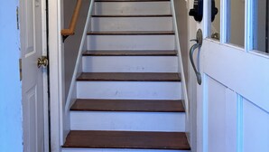 Un escalier avec des marches en bois et des murs blancs, menant à une porte dotée d’une poignée en laiton.