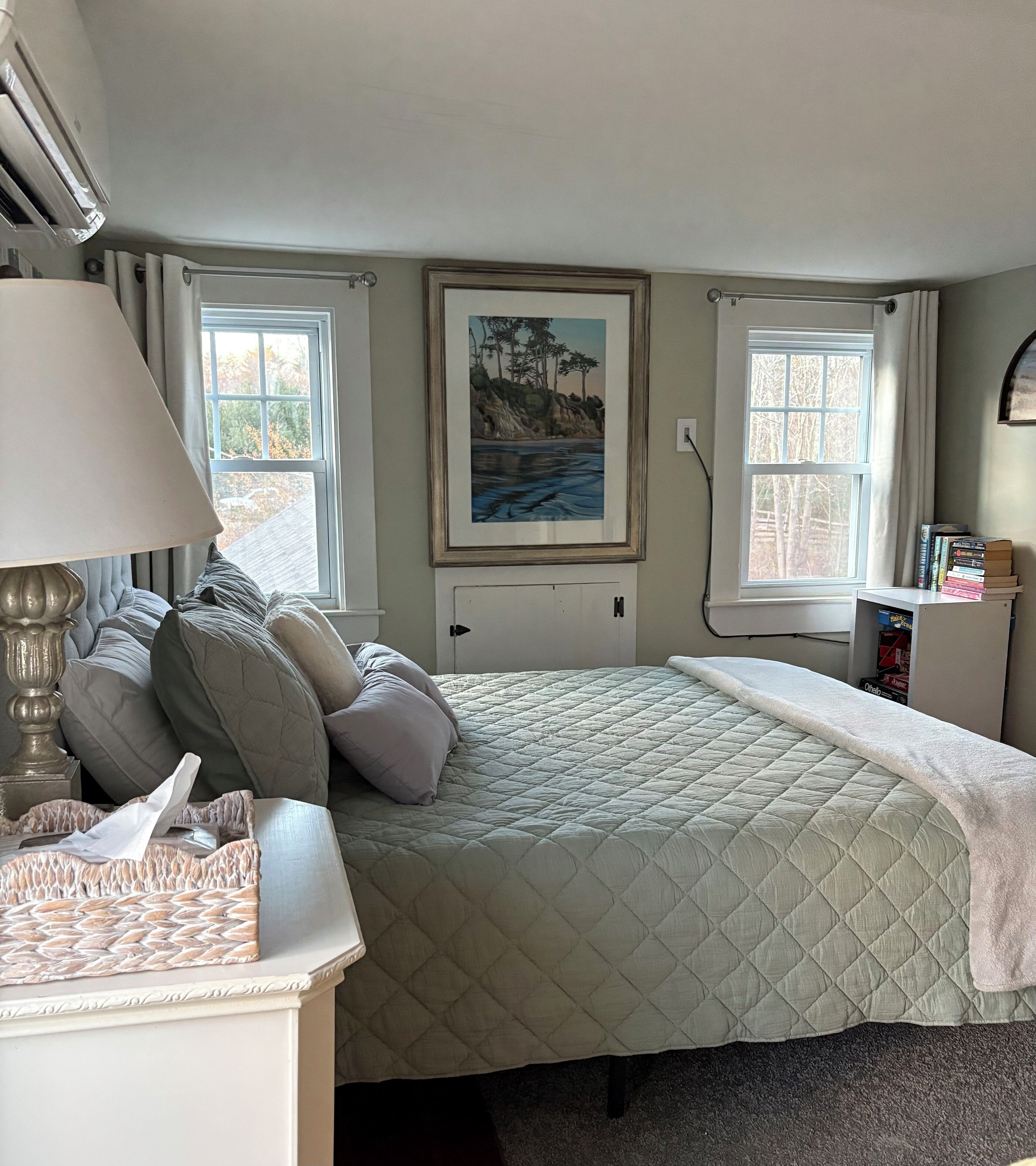 A bedroom with a bed, a bedside table, a lamp, a painting on the wall, and two windows.
