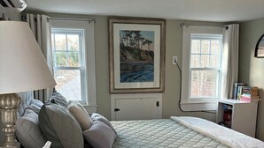 Une chambre à coucher avec un lit, une table de chevet, une lampe, un tableau au mur et deux fenêtres.