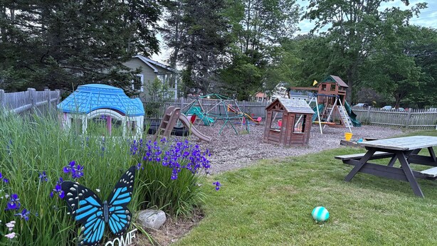Un jardin avec un espace de jeux, une table de pique-nique et une pancarte « BIENVENUE » au milieu des fleurs.