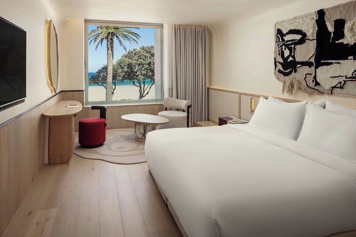 A hotel room with a large bed, a TV, a window with a view of palm trees and the sea, a small table, and a red ottoman.