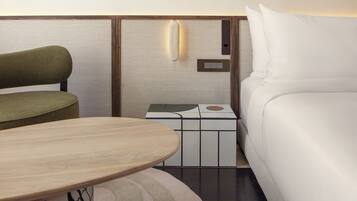 A modern hotel room with a bed, a green upholstered chair, a wooden table, and a nightstand with a lamp.