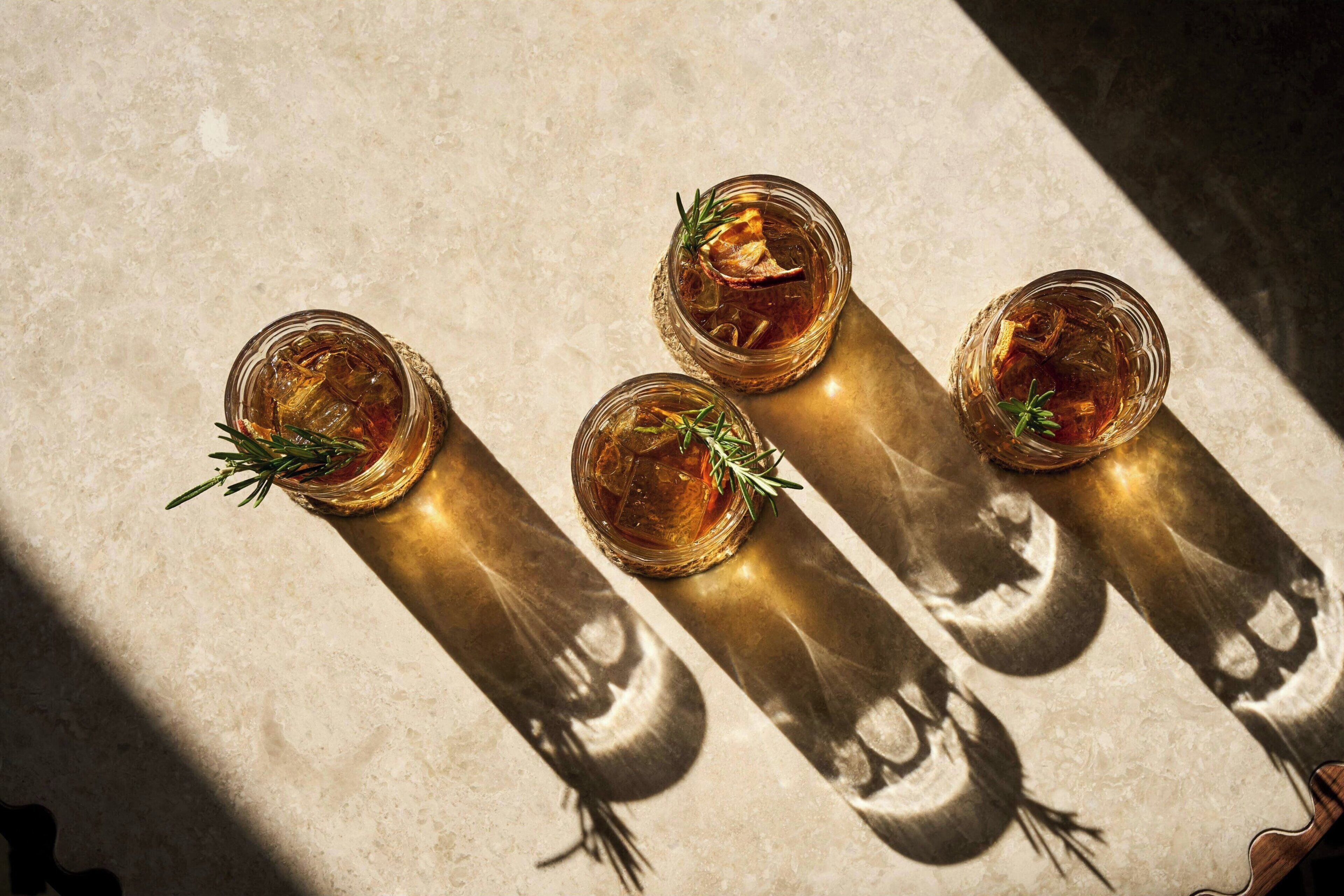 Three glasses of amber liquid with rosemary sprigs, placed on a textured surface with shadows.