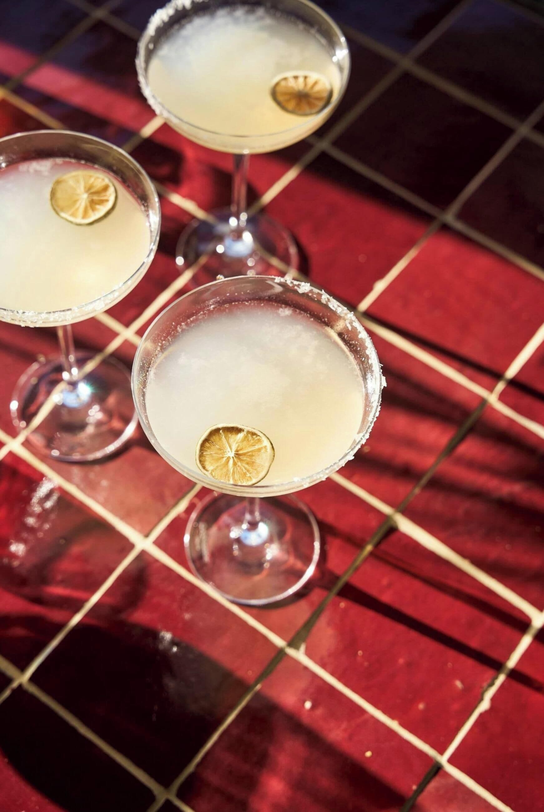 Trois verres à martini, décorés d’une tranche de citron, posés sur une surface recouverte de carreaux rouges.
