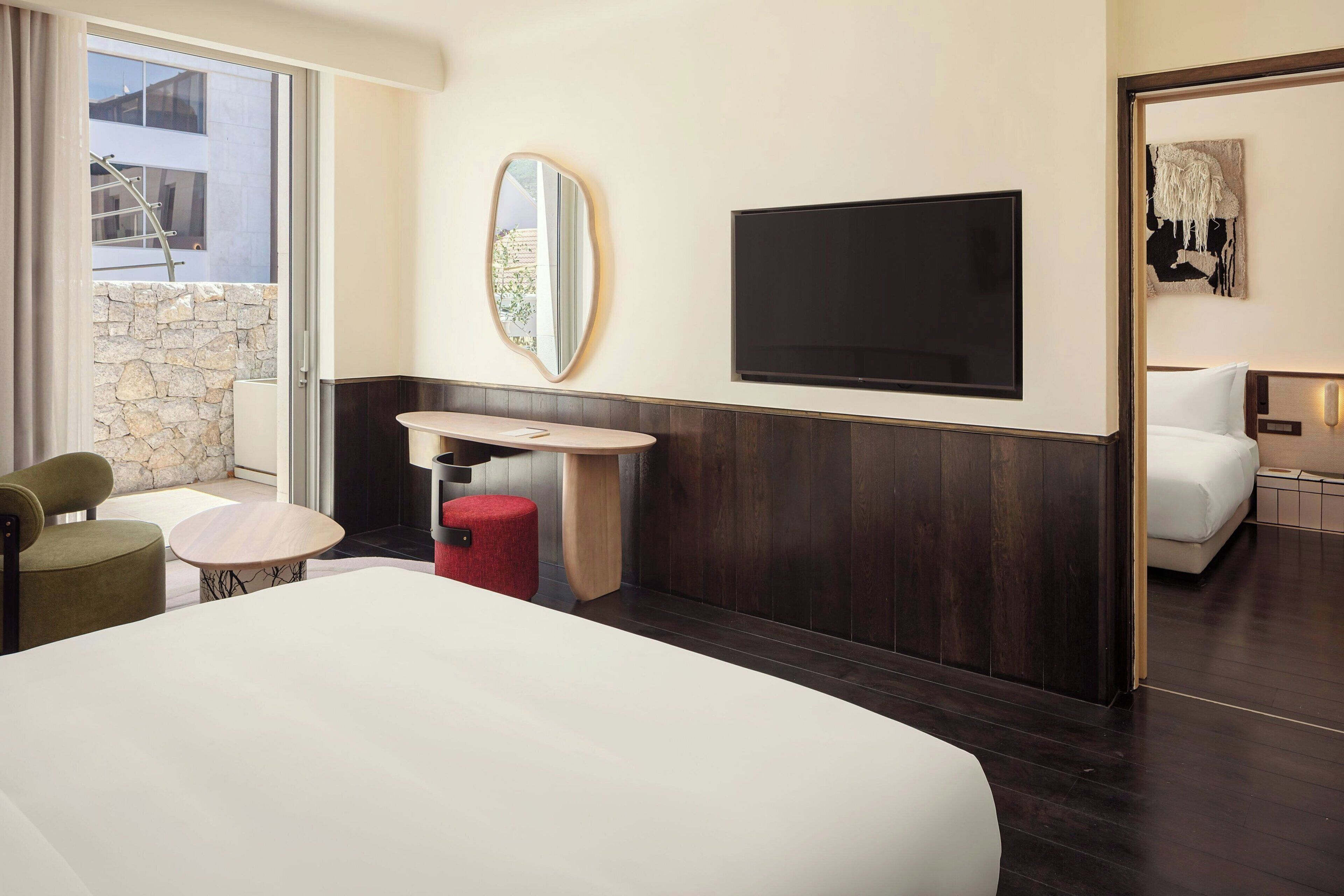 A hotel room with a large bed, a TV mounted on the wall, a small round table, and a red stool.