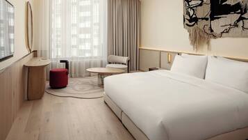 A modern hotel room with a large bed, a small round table, a red ottoman, and a large window with sheer curtains.