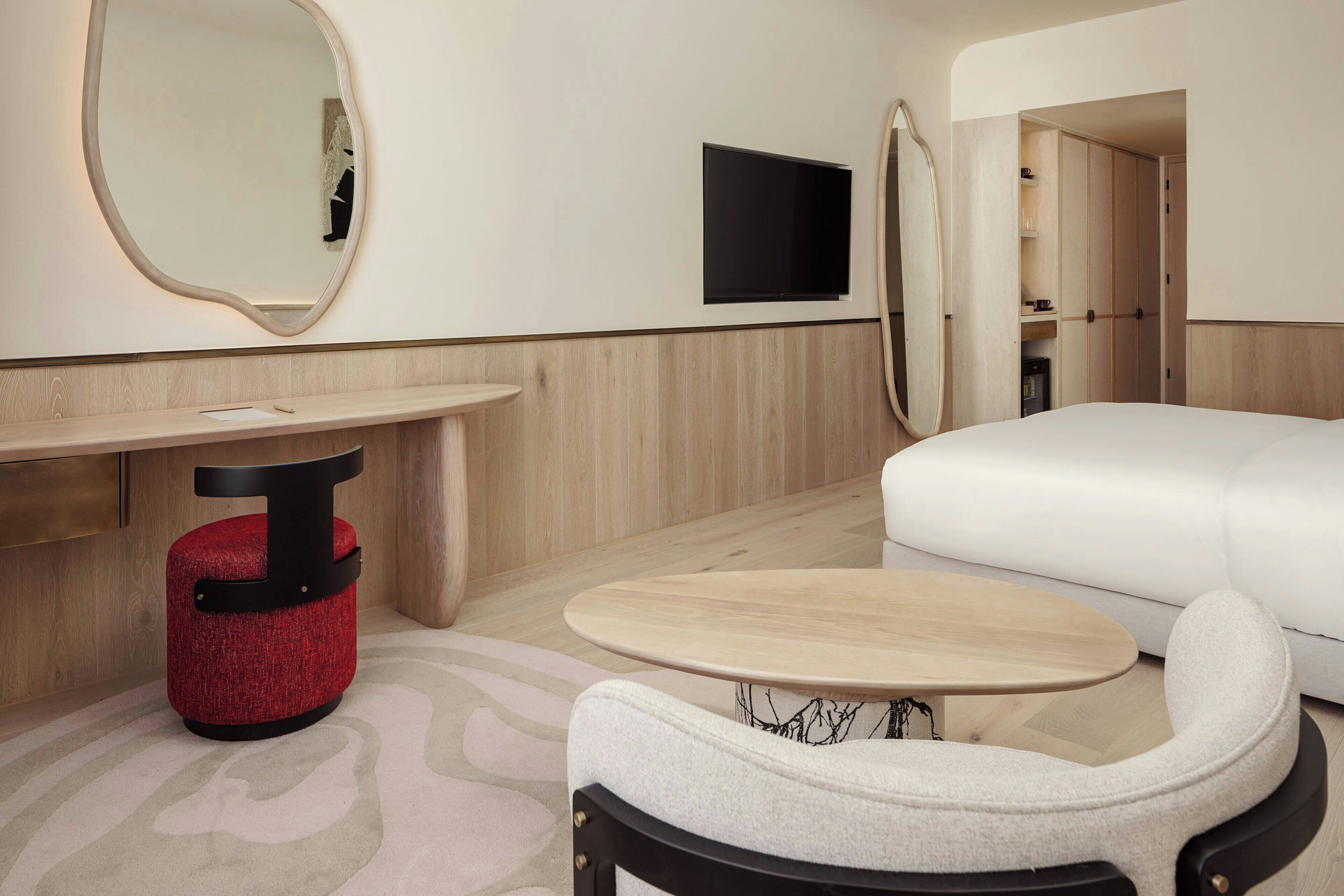 A modern hotel room with a sofa, a small round table, a red ottoman, a mirror, and a flat-screen TV mounted on the wall.