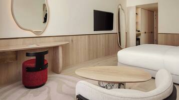A modern hotel room with a sofa, a small round table, a red ottoman, a mirror, and a flat-screen TV mounted on the wall.