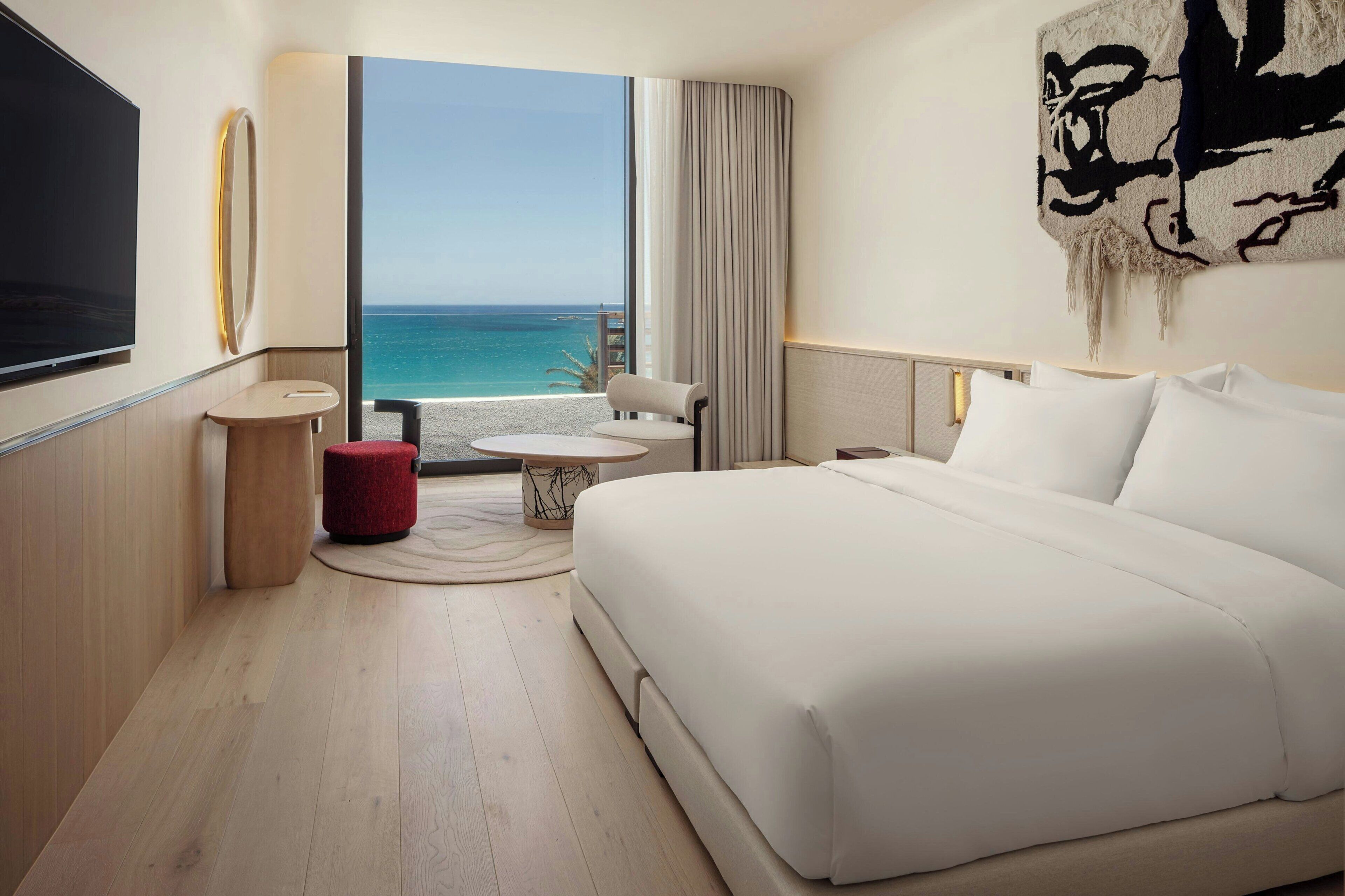 A hotel room with a large bed, a TV, a small table, and a view of the ocean.