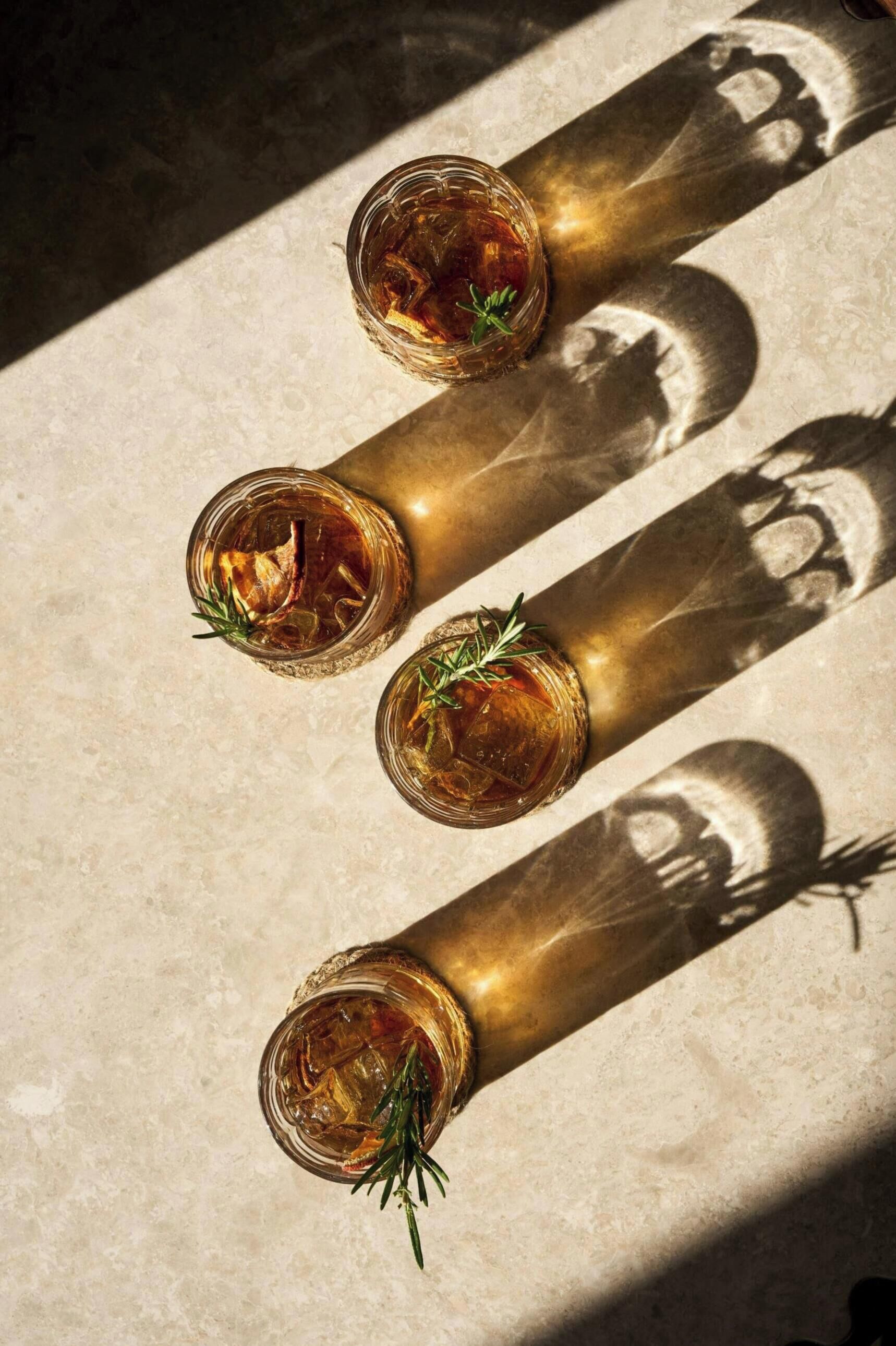 Three glasses of amber liquid with rosemary garnish on a textured surface, cast in shadows.