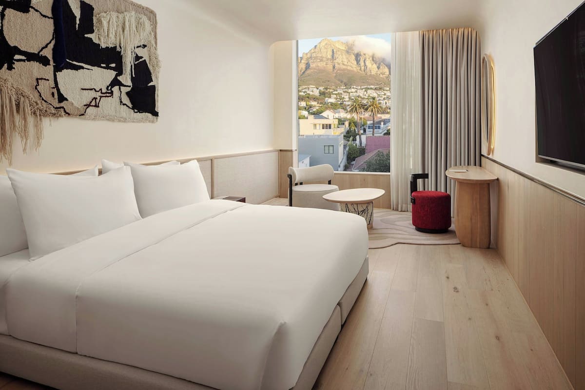 A hotel room with a large bed, a small round table, a red ottoman, and a view of a mountain and town through the window.