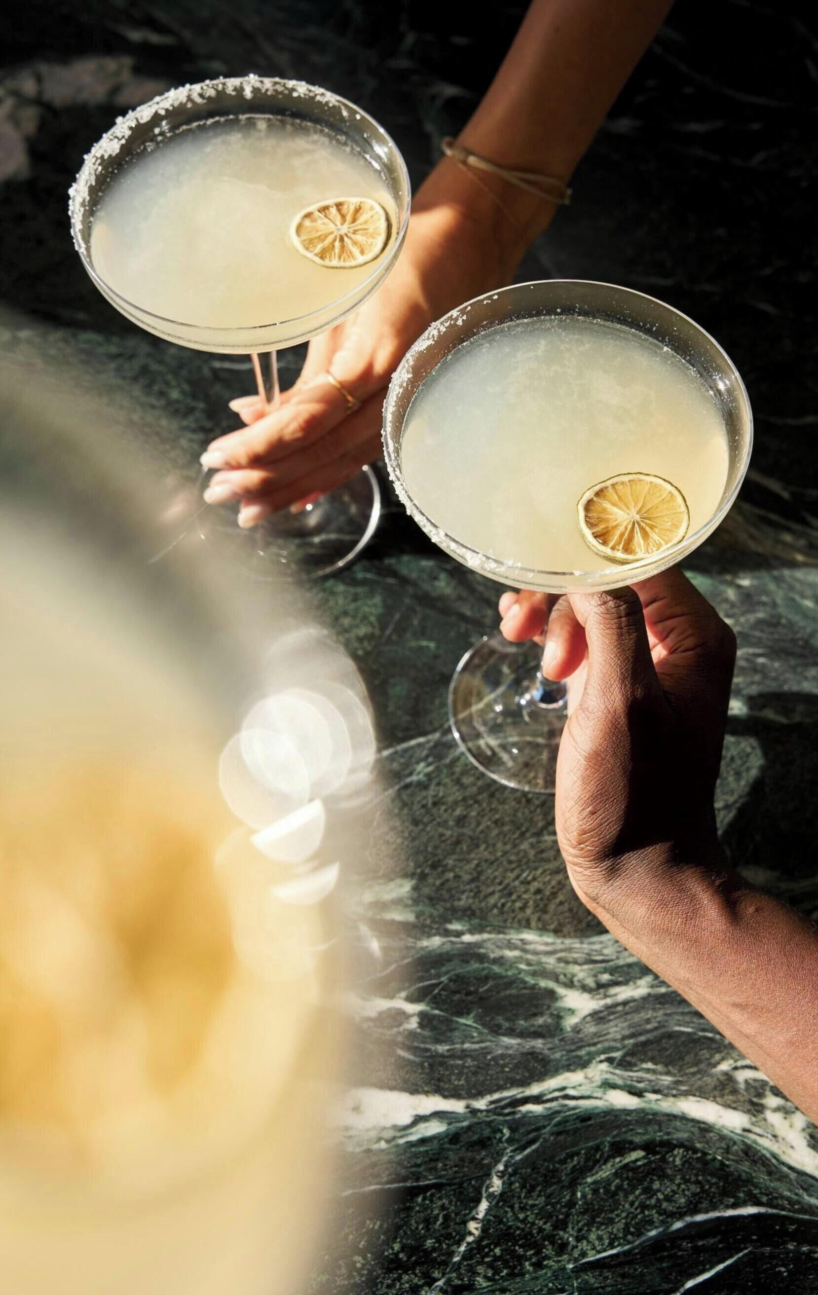 Two people holding margaritas with a lime garnish, on a marble surface.