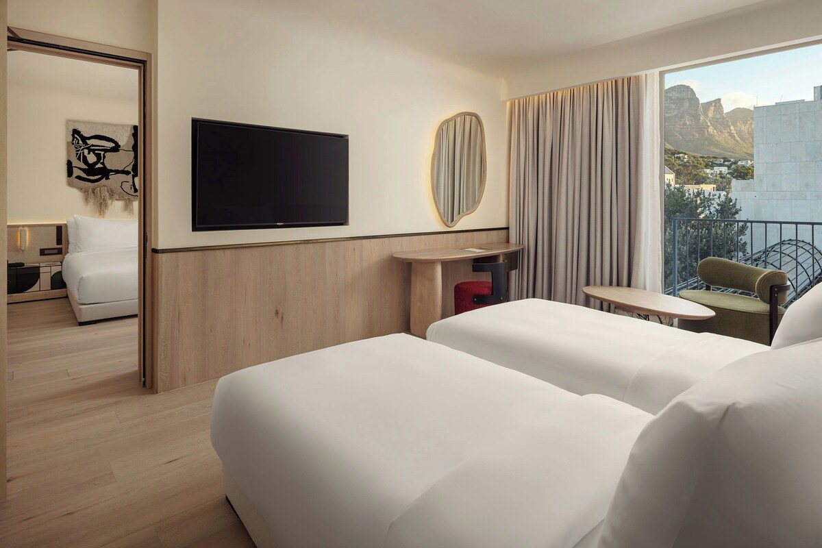A hotel room with two beds, a TV, a mirror, a small table, and a balcony with a view of mountains.