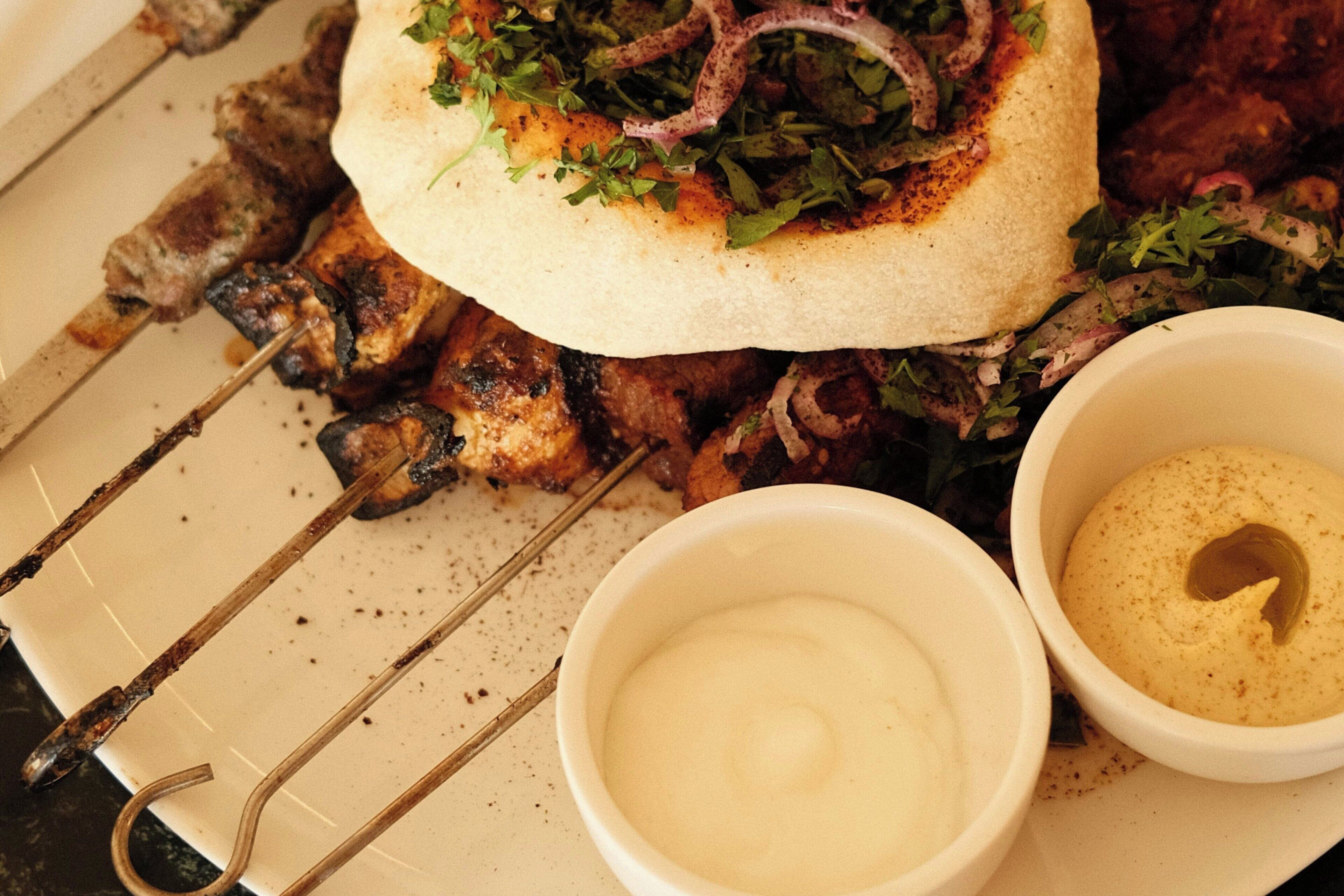 Un piatto con spiedini di carne alla griglia, una piadina farcita con verdure e cipolle, e due piccole ciotole di salsa.