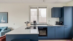 A modern kitchen with a central island, a built-in dishwasher, and a window with blinds. - WW | Dijkhuys Buitenbubbelbad (Opperdoes)