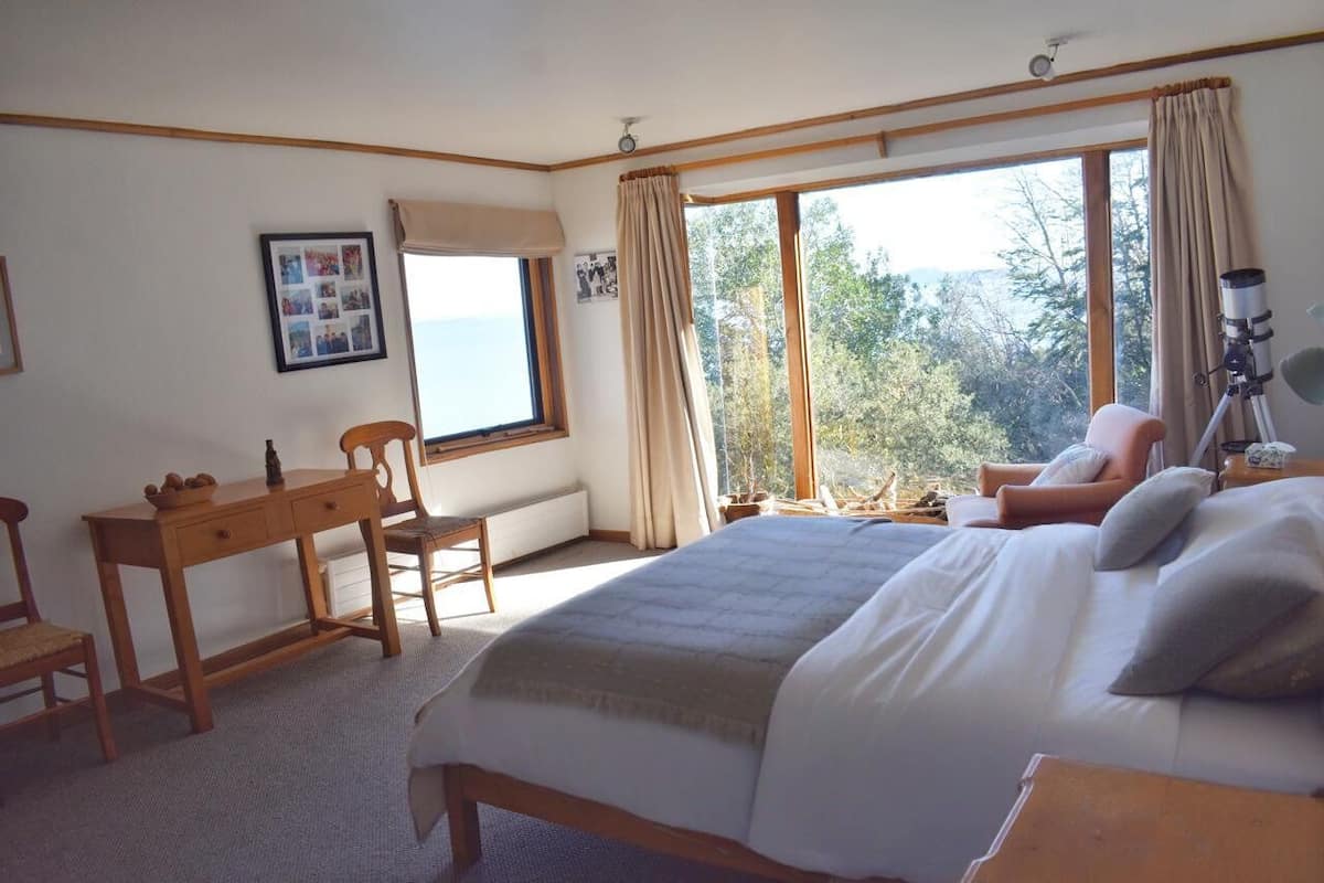 A bedroom with a large bed, a desk, a chair, and a window with a view of trees.