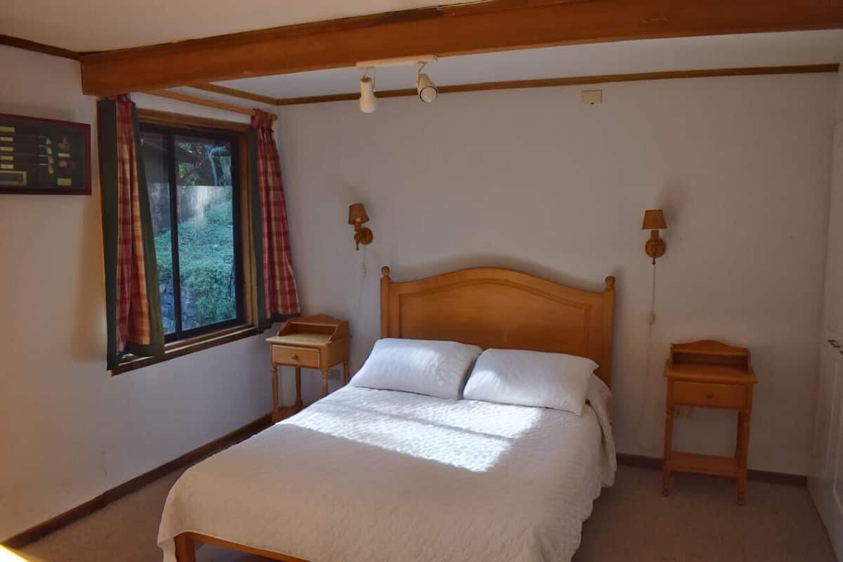 A bedroom with a wooden bed, two bedside tables, a window with curtains, and a wall-mounted lamp.