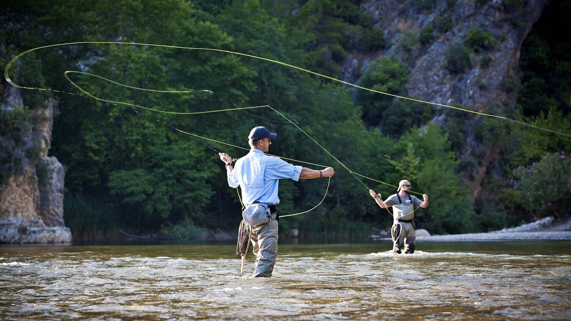 Two individuals fly fishing in a river with a rocky cliff in the background.