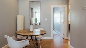 Un área de comedor con una mesa de madera redonda, dos sillas blancas, un espejo grande y vista a un pasillo.