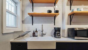 Una cocina con un lavamanos blanco, armarios negros, un estante de madera, una cafetera y un microondas.