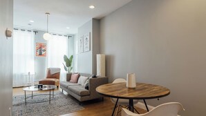 Una sala de estar moderna con un sofá, una mesa de centro y sillas. Hay un ventanal con cortinas, una planta y un cartel en la pared.