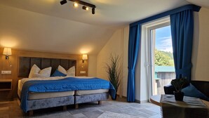 A bedroom with a large bed, a nightstand, a lamp, and a view of the outdoors through a window with blue curtains.