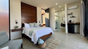 Une chambre moderne avec un grand lit, une table de chevet, un petit bureau, un espace douche et une salle de bain équipée d’un lavabo et d’un miroir.