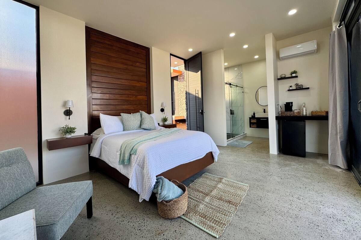 Quarto moderno com uma cama grande, uma mesinha de cabeceira, uma pequena escrivaninha, área de chuveiro e banheiro com pia e espelho.