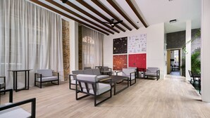 Een ruime kamer met een modern ontwerp, met een houten plafond, een accentmuur van steen en een groot raam met transparante gordijnen. Er zijn meerdere zithoeken met tafels en stoelen, en een muur met diverse infobrochures.