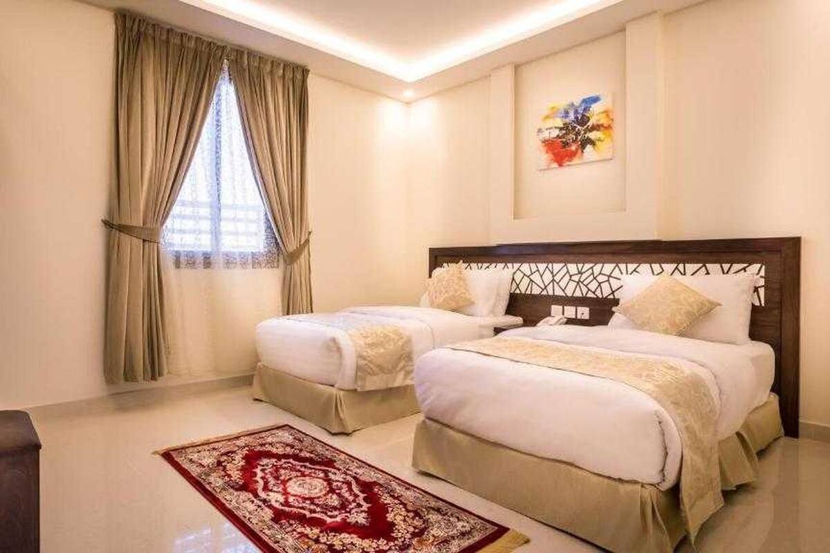 A hotel room with two beds, a wooden headboard, a red patterned rug, a window with curtains, and a painting on the wall.