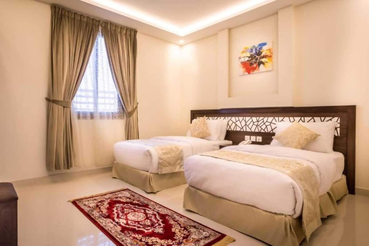 A hotel room with two beds, a wooden desk, a red patterned rug, and a window with curtains.