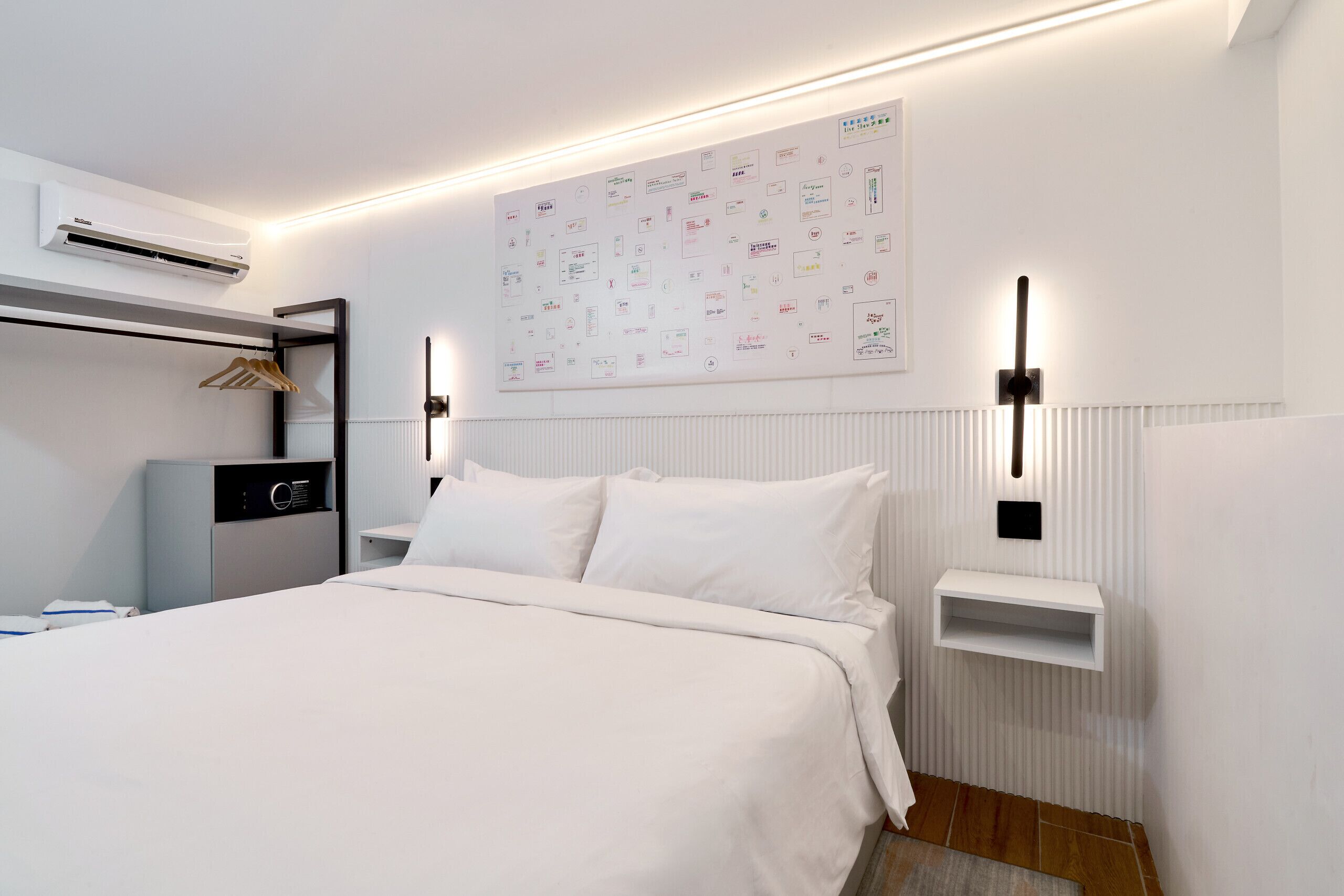 Une chambre d’hôtel moderne avec un lit bien fait, un climatiseur fixé au mur, une table de chevet avec une lampe et un tableau d’affichage où sont accrochées diverses notes.
