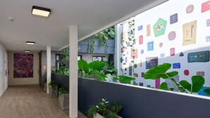A hallway with a modern design, featuring a long corridor with a wooden floor, a large abstract painting on the left wall, and a series of planters with greenery along the right wall. - Exclusively designed room CG15 (Ciudad de Panamá)