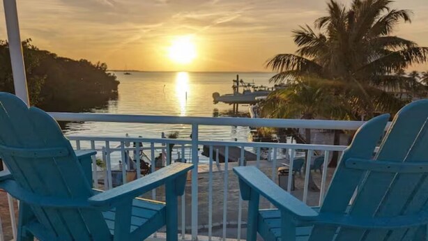 A balcony with two blue Adirondack chairs facing a sunset over water. - Charming 2-bedroom campground in Key Largo with AC (Key Largo)