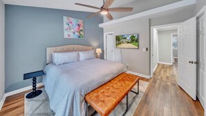A bedroom with a bed, a bench, a ceiling fan, a painting, and a TV on the wall.
