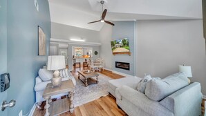 A living room with a sofa, coffee table, and TV. There's a fireplace and a dining area in the background.