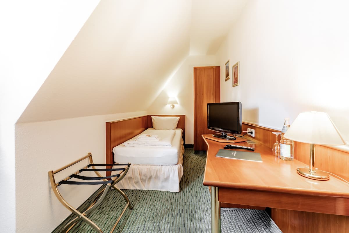 Una habitación de hotel pequeña y ordenada con una cama individual, un escritorio con computadora, una silla y una lámpara.