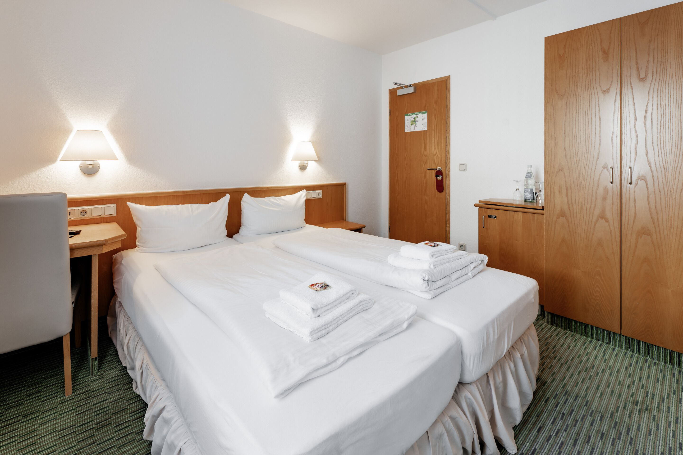 A hotel room with two beds, a desk, a chair, a wardrobe, and two wall-mounted lamps.