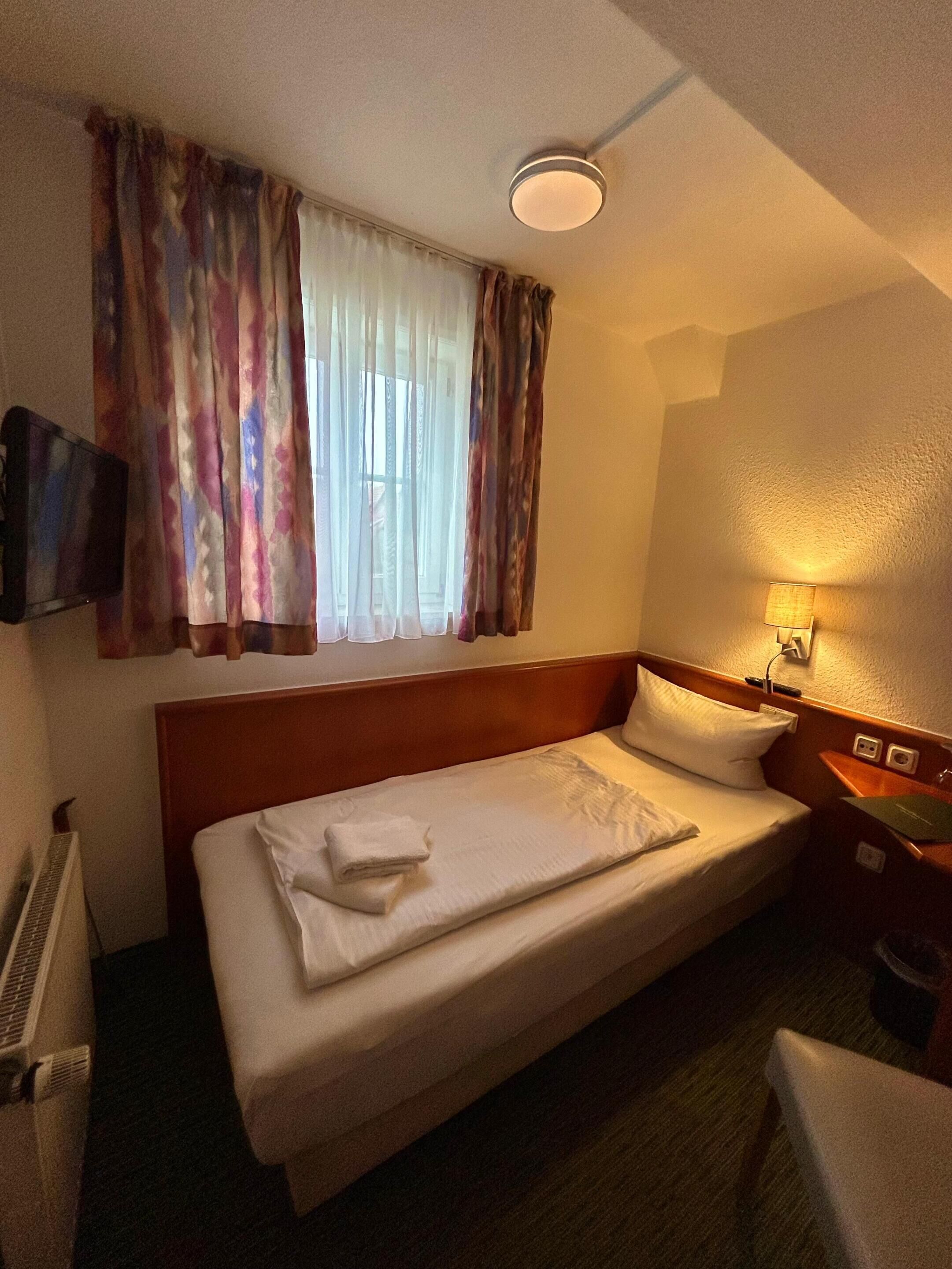 Ein Einzelbett mit Holz Kopfteil, ein Nachttisch mit Lampe, ein Flachbildfernseher an der Wand und ein Fenster mit durchsichtigen Vorhängen.