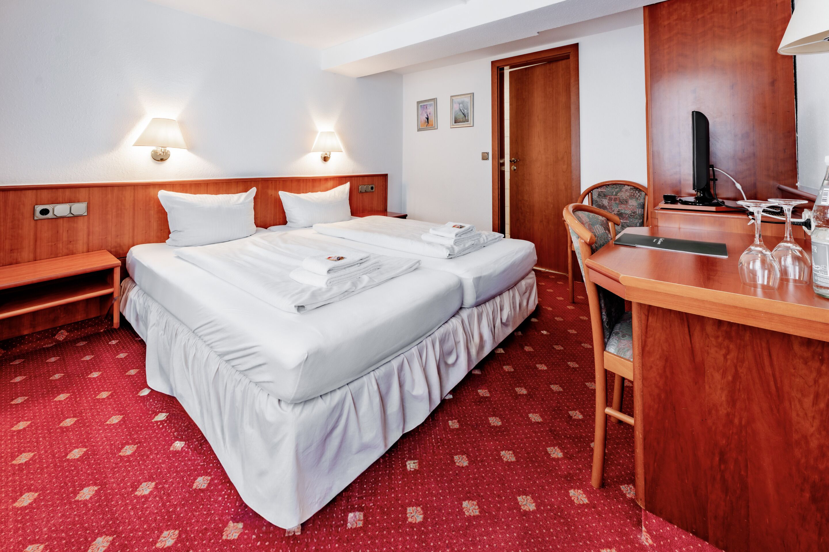 Ein Hotelzimmer mit einem Bett, Nachttischen, einem Schreibtisch mit Stuhl, einem Fernseher und einem roten Teppichboden.