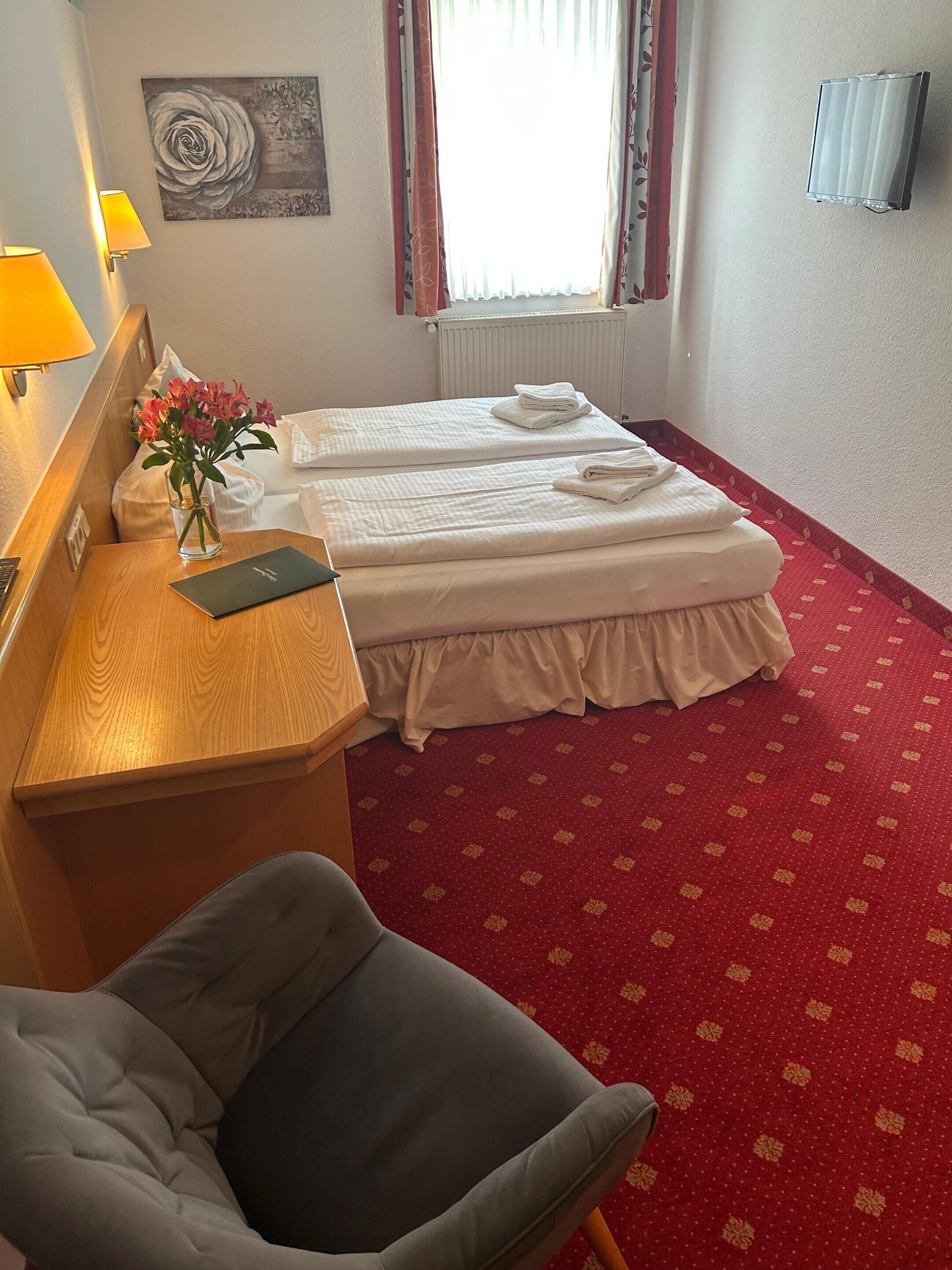Habitación de hotel con dos camas, un escritorio con lámpara, una silla, un florero con flores y un cuadro en la pared.