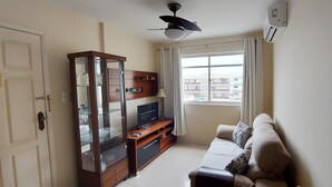A living room with a sofa, a wooden cabinet with glass doors, a television, and a window with curtains. - COOND0115 - Edifício Regina (Salvador)