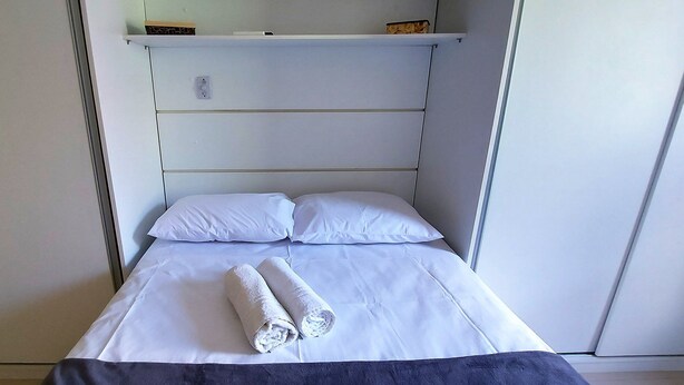 A single bed with white linens and two pillows, a rolled towel on the bed, and a shelf above the bed. - COOND0115 - Edifício Regina (Salvador)
