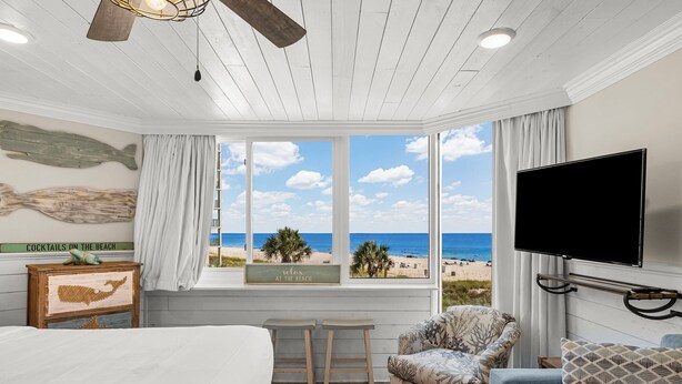 Hotelkamer met een groot raam met uitzicht op het strand, een bed, een televisie en een zithoek met een gestreept fauteuil en poef.