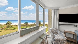 Een kamer met een groot raam dat uitkijkt op een strand, een flatscreen televisie aan de muur en een gestreept bankje.