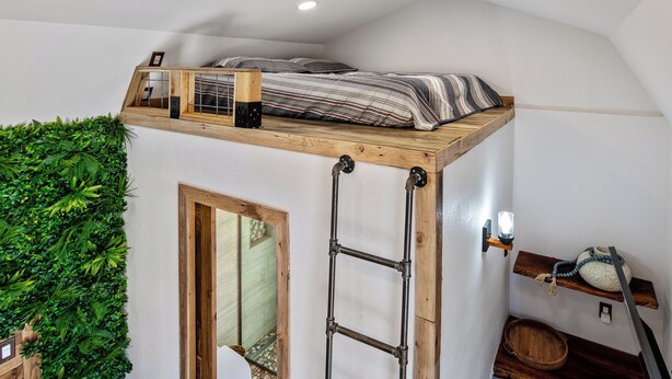 Dormitorio estilo loft con cama sobre plataforma de madera, escalera, pared verde y un estante pequeño con lámpara y objetos decorativos.