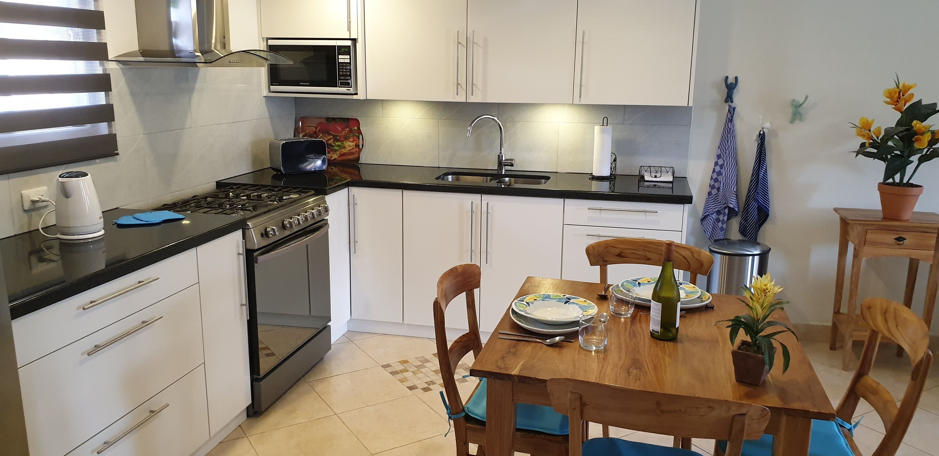 Una cocina moderna con armarios blancos, encimera negra, microondas, tostadora y fregadero. Hay una mesa de comedor con sillas, una botella de vino y una planta en maceta.