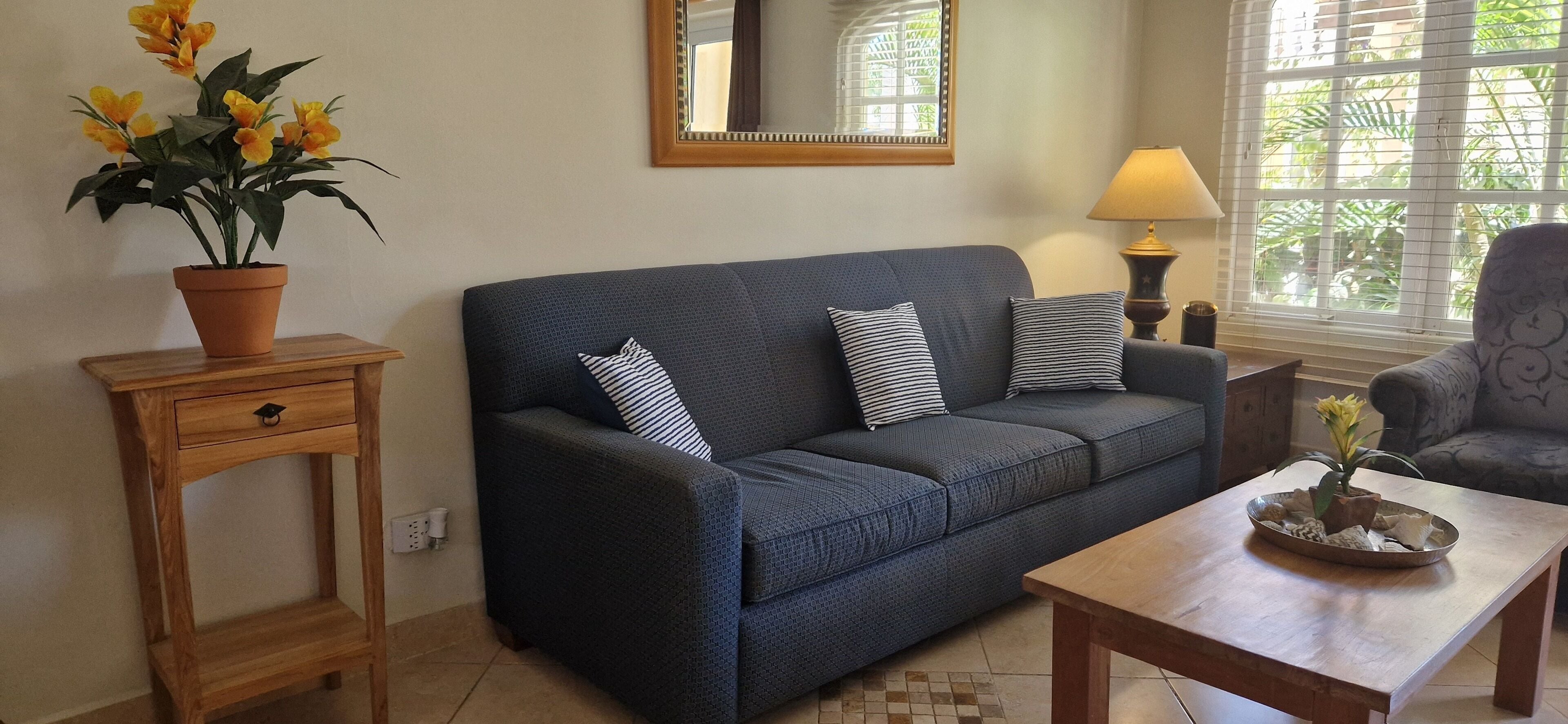 A living room with a dark blue sofa, two striped pillows, a wooden side table with a potted plant, a wooden coffee table with decorative items, a lamp, and a window with blinds.
