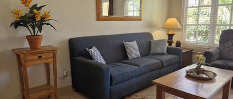 A living room with a dark blue sofa, two striped pillows, a wooden side table with a potted plant, a wooden coffee table with decorative items, a lamp, and a window with blinds.